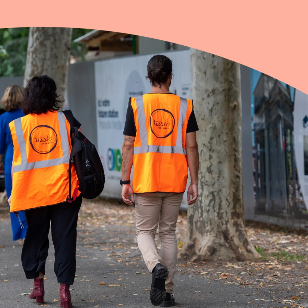 Deux personnes de dos avec un gilet orange avec un logo Tisséo Ingenierie qui marchent près du chantier de la ligne C de Toulouse